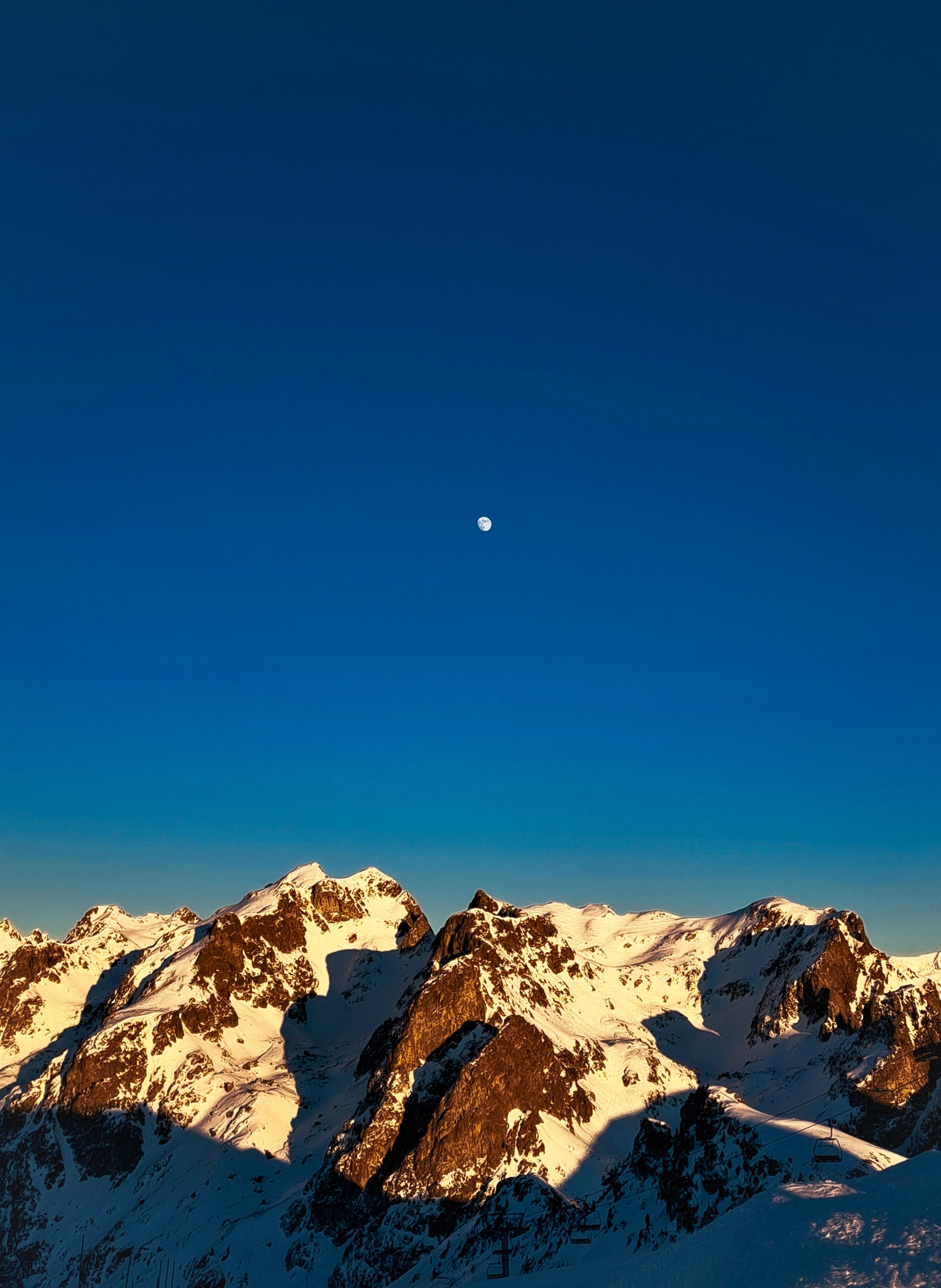Lune X Montage -- Fin de journée de ski sur le domaine de Chamrousse, où la Lune émerge tandis que les montagnes prennent des teintes chaudes. C'est un instant particulier où le ciel se pare de nuances douces et la nature semble s'endormir paisiblement après une journée d'activités.