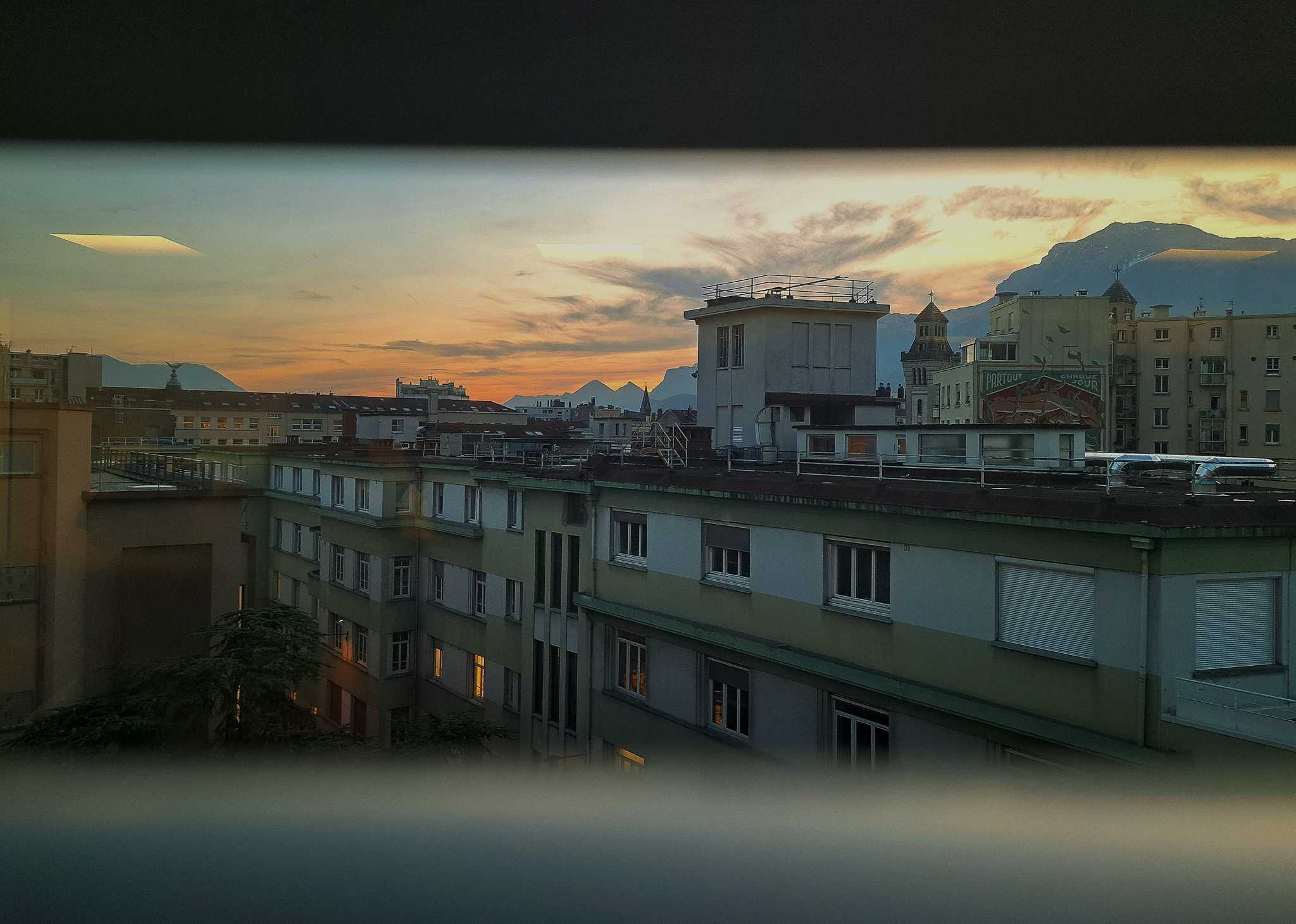 L'avant nuit -- Ici, à travers cette fenêtre, se dessinent les toits de Grenoble, éclairés par la lumière douce du soleil couchant. C'est un moment où la ville semble prendre une teinte différente, presque irréelle, sous cette lumière magique qui transforme chaque coin de rue en une scène d'une beauté éphémère.