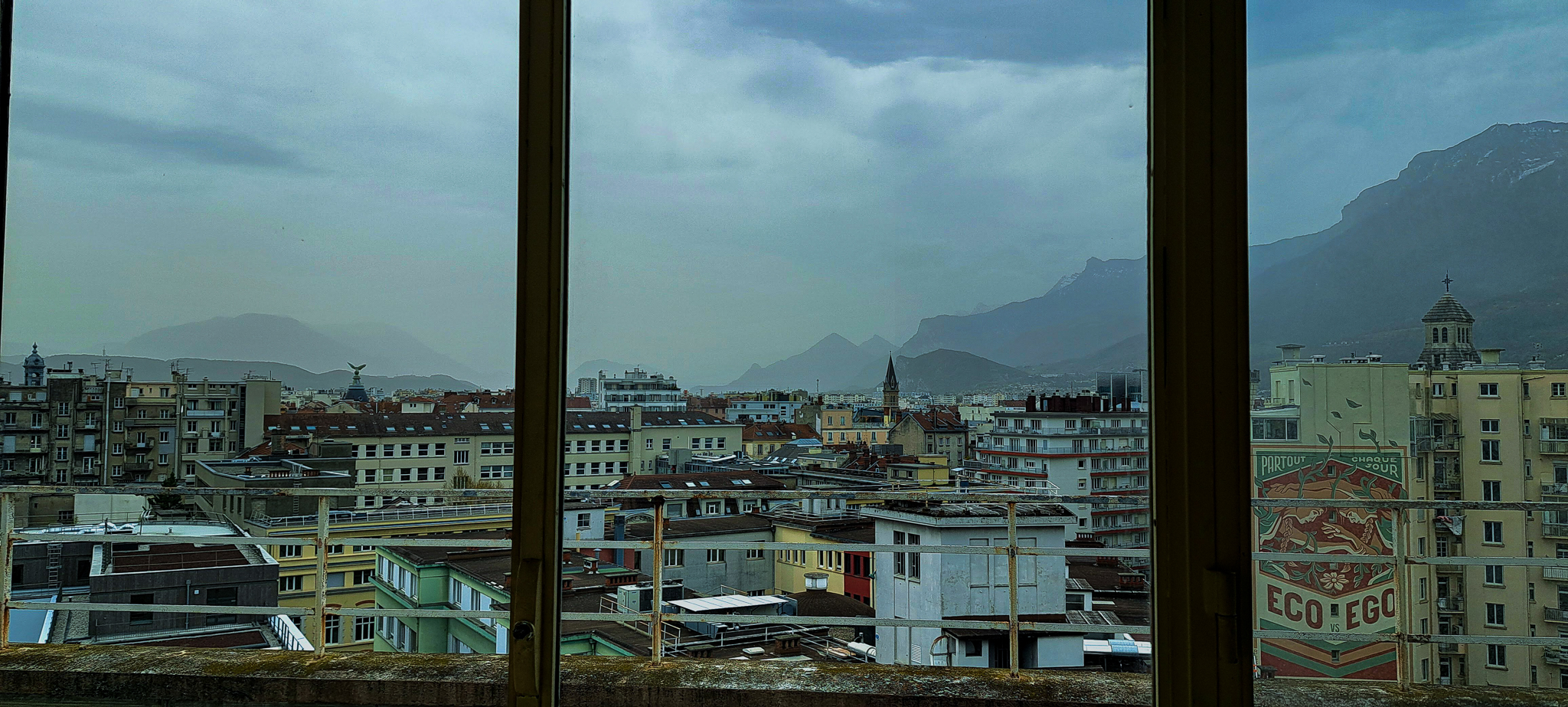 Sous un ciel maussade, la vie s'anime sous les toits de Grenoble. Malgré la météo morose, l'activité incessante de la ville contraste avec l'atmosphère calme et contemplative qui règne au-dessus.
